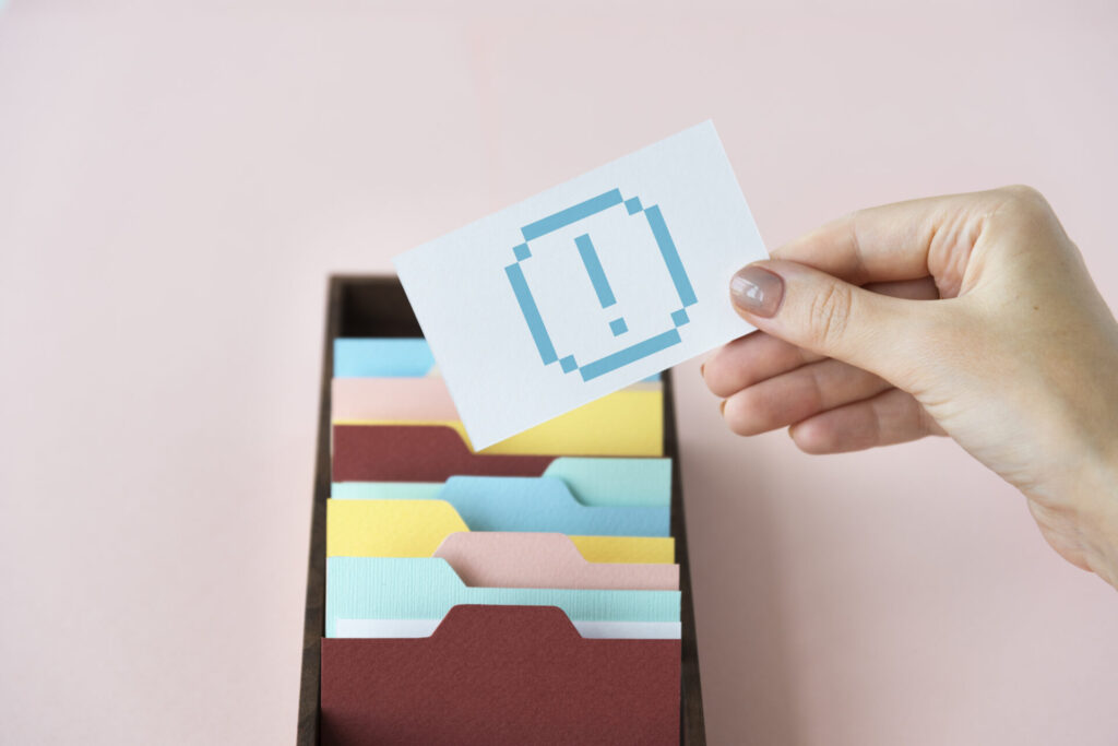 代替テキスト案: ファイルボックスから「!」のアイコンが描かれたカードを取り出す手のクローズアップ。会員登録しない場合の注意点やデメリットを象徴している。