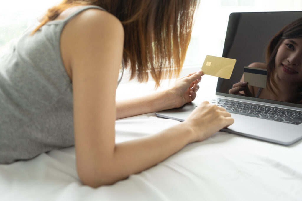 代替テキスト案: ベッドの上でノートパソコンを使い、クレジットカードを手に持ってオンラインショッピングをしている女性。ユニクロのオンラインストアでの購入や特典活用シーンを表している。