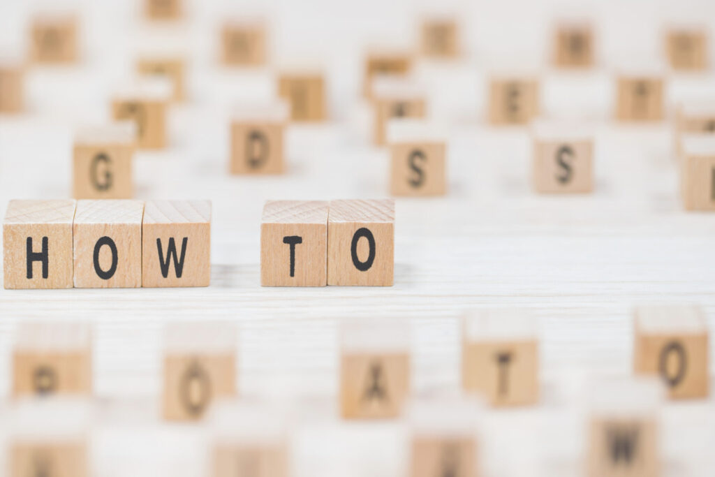 木製のサイコロ状のブロックが散らばっており、手前には「HOW TO」と書かれたブロックが並べられています。他のブロックには様々なアルファベットが書かれており、ハウツー記事や攻略法を視覚的に表現しています。